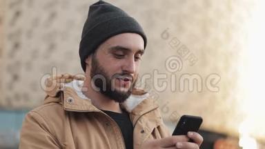 快乐的年轻胡须男在购物中心附近的街道上使用智能手机。 他穿着一件秋装和一顶针织帽子